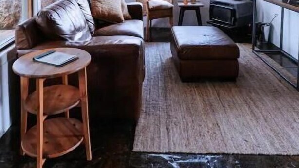 Ein gemütliches Wohnzimmer mit einem Ledersofa, einem Holztisch und einem Fernseher. Ein großes Fenster sorgt für natürliches Licht und einen Blick ins Freie.