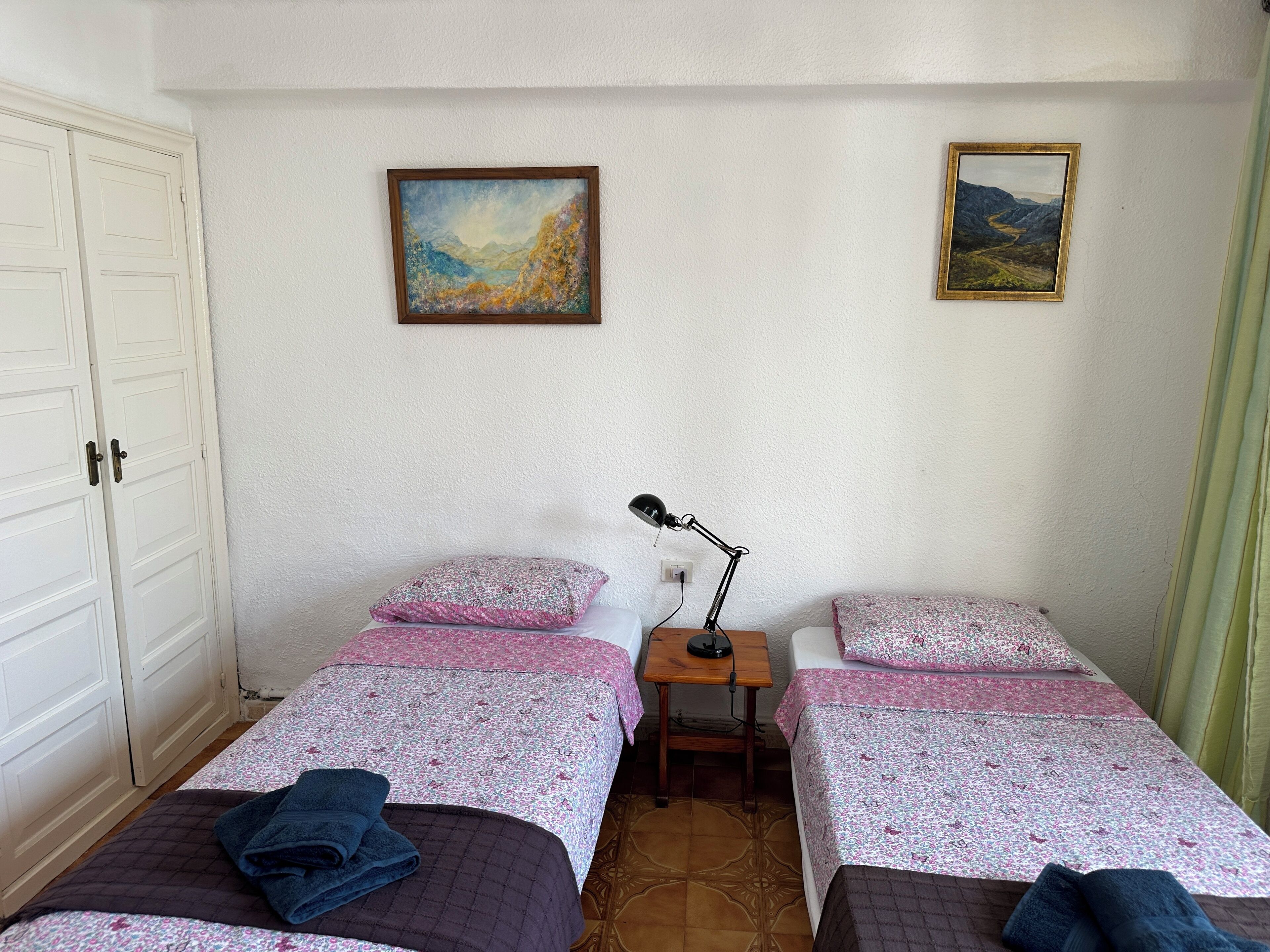 A room with two beds, each with a floral bedspread and pillows, a small table with a lamp, and two framed pictures on the wall.