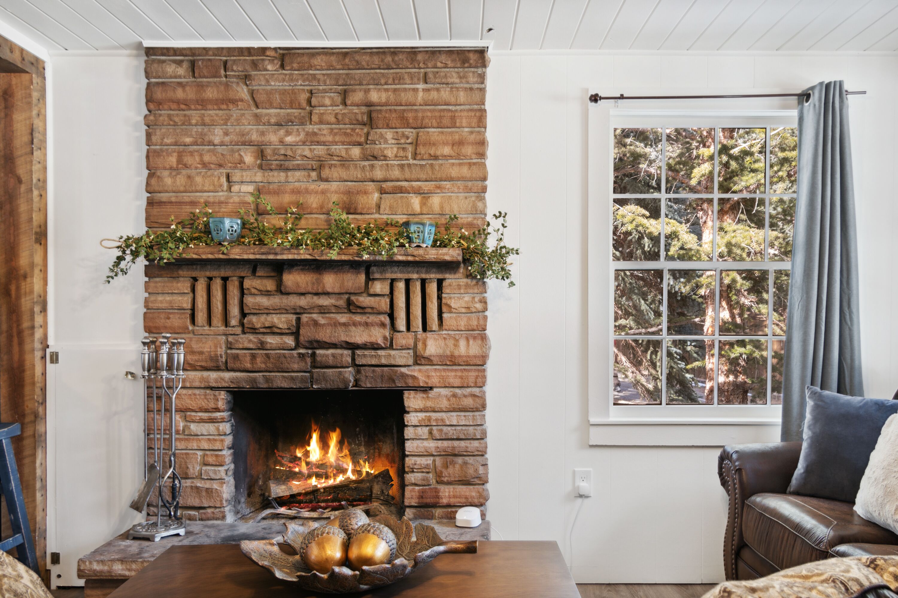 A cozy living room with a stone fireplace, a window with a view of trees, and a comfortable seating area.