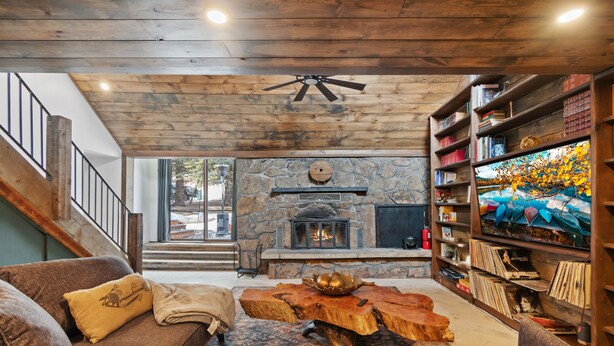 A cozy living room with a stone fireplace, wooden ceiling, and a bookshelf filled with books. - Ferncliff Lodge (Allenspark)