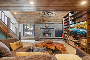 A cozy living room with a stone fireplace, wooden ceiling, and a bookshelf filled with books. - Ferncliff Lodge (Allenspark)