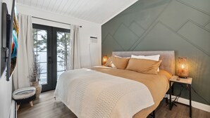 A bedroom with a bed, a bedside table with a lamp, a window with curtains, and a door leading outside. - Ferncliff Lodge (Allenspark)