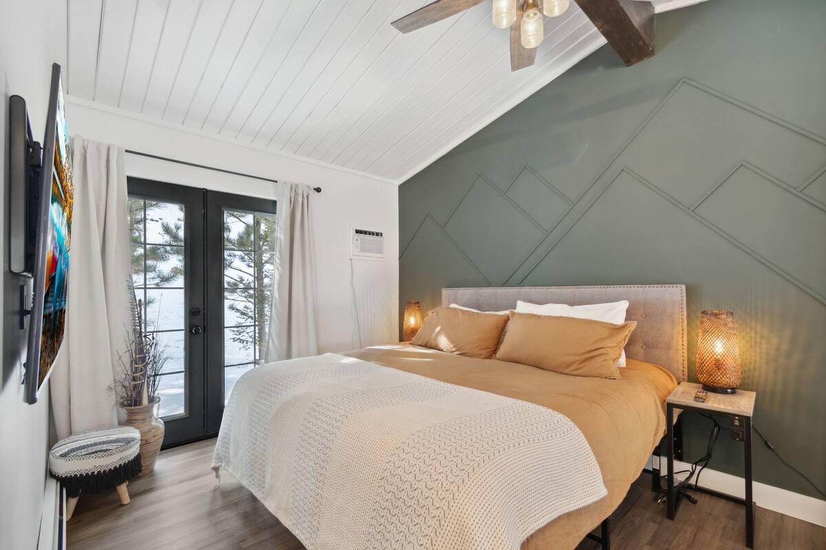 A bedroom with a bed, a bedside table with a lamp, a window with curtains, and a door leading outside.
