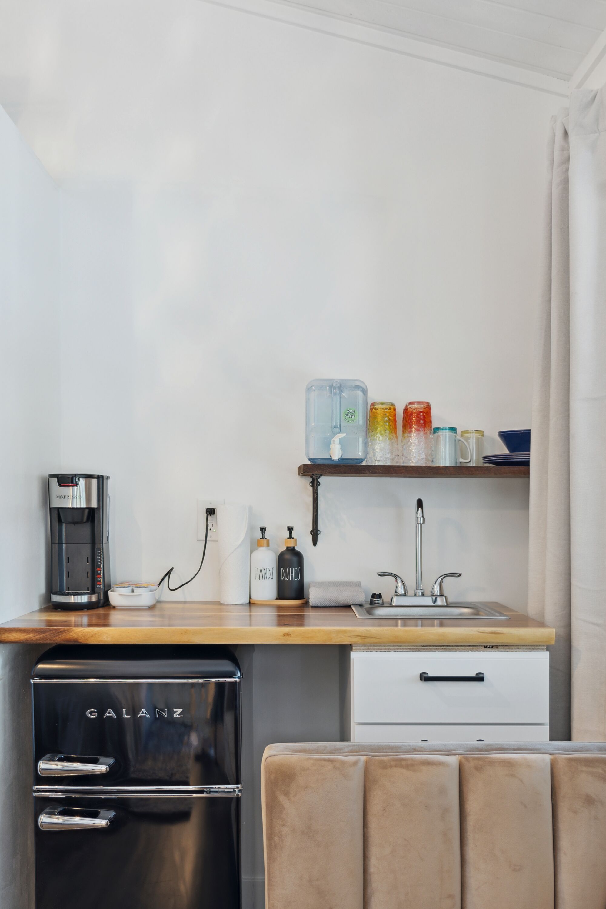 A compact kitchenette with a coffee maker, a mini-fridge, a sink, and a shelf with various containers.