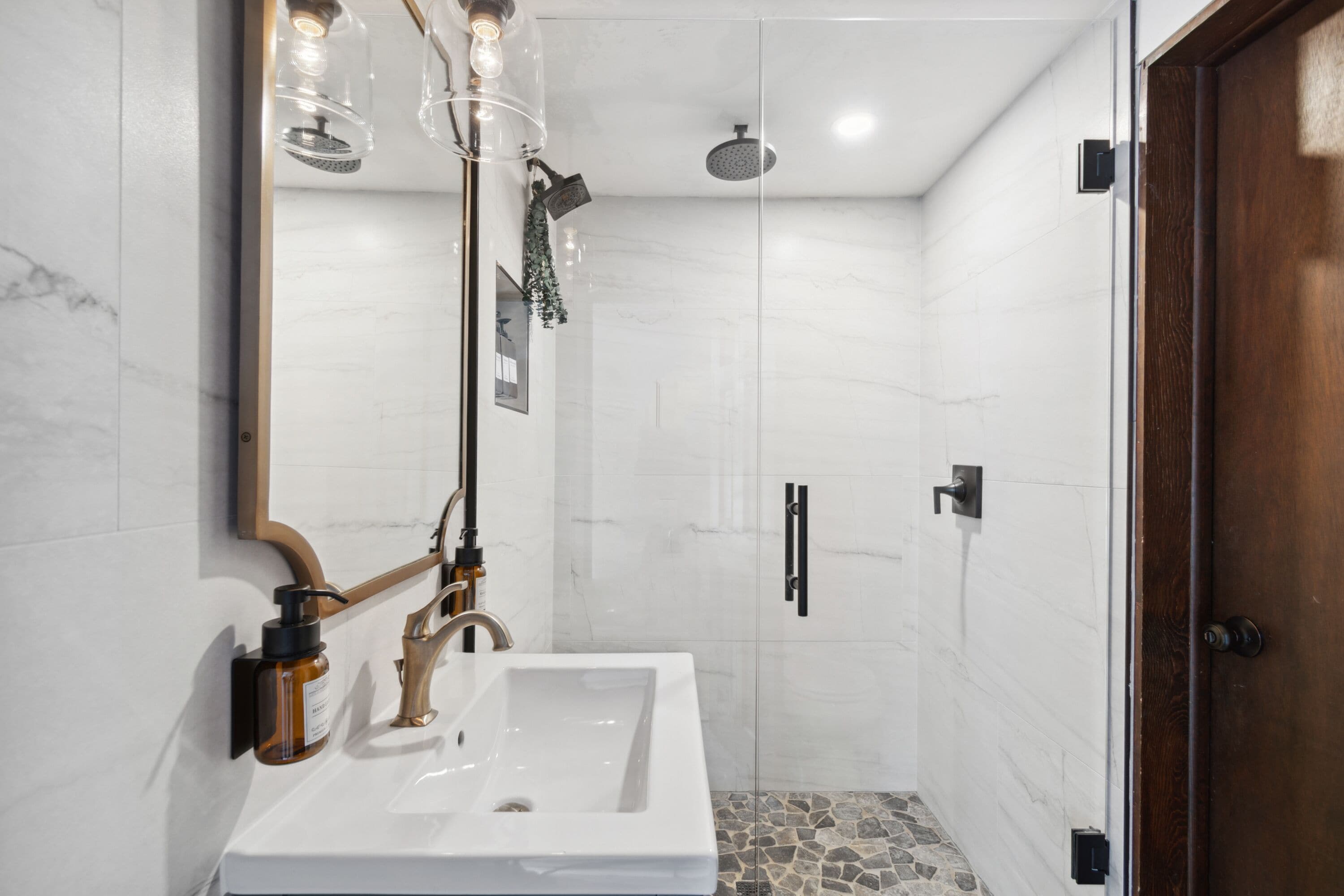 Un baño moderno con un lavabo blanco, un espejo, una ducha con cabezal de ducha manual y una puerta de madera.