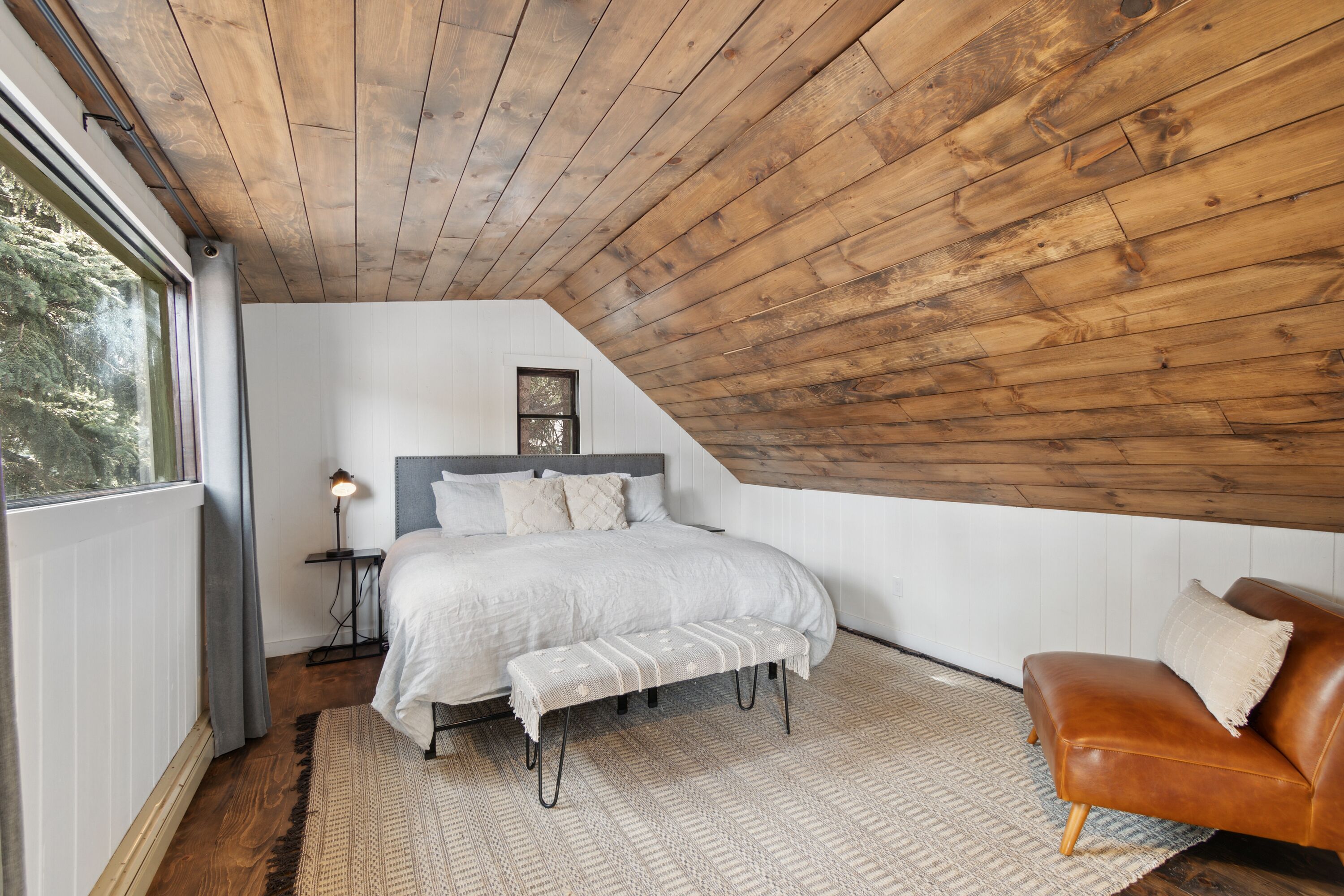 Un dormitorio con techo inclinado, una cama con cabecera, un banco, un sillón de cuero marrón y una mesita de noche con lámpara.