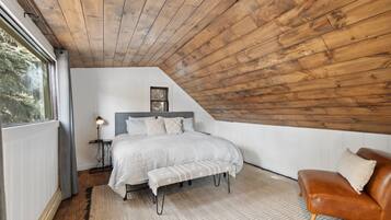 Un dormitorio con techo inclinado, una cama con cabecera, un banco, un sillón de cuero marrón y una mesita de noche con lámpara.