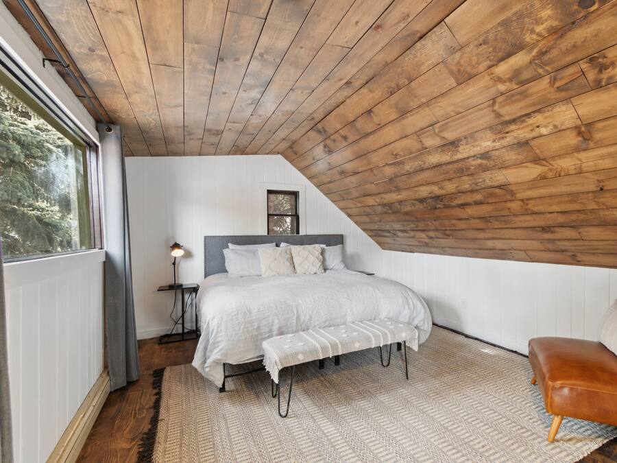 Un dormitorio con techo inclinado, una cama con cabecera, un banco, un sillón de cuero marrón y una mesita de noche con lámpara.