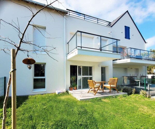 A modern two-story house with a balcony, a patio with outdoor furniture, and a well-maintained lawn.