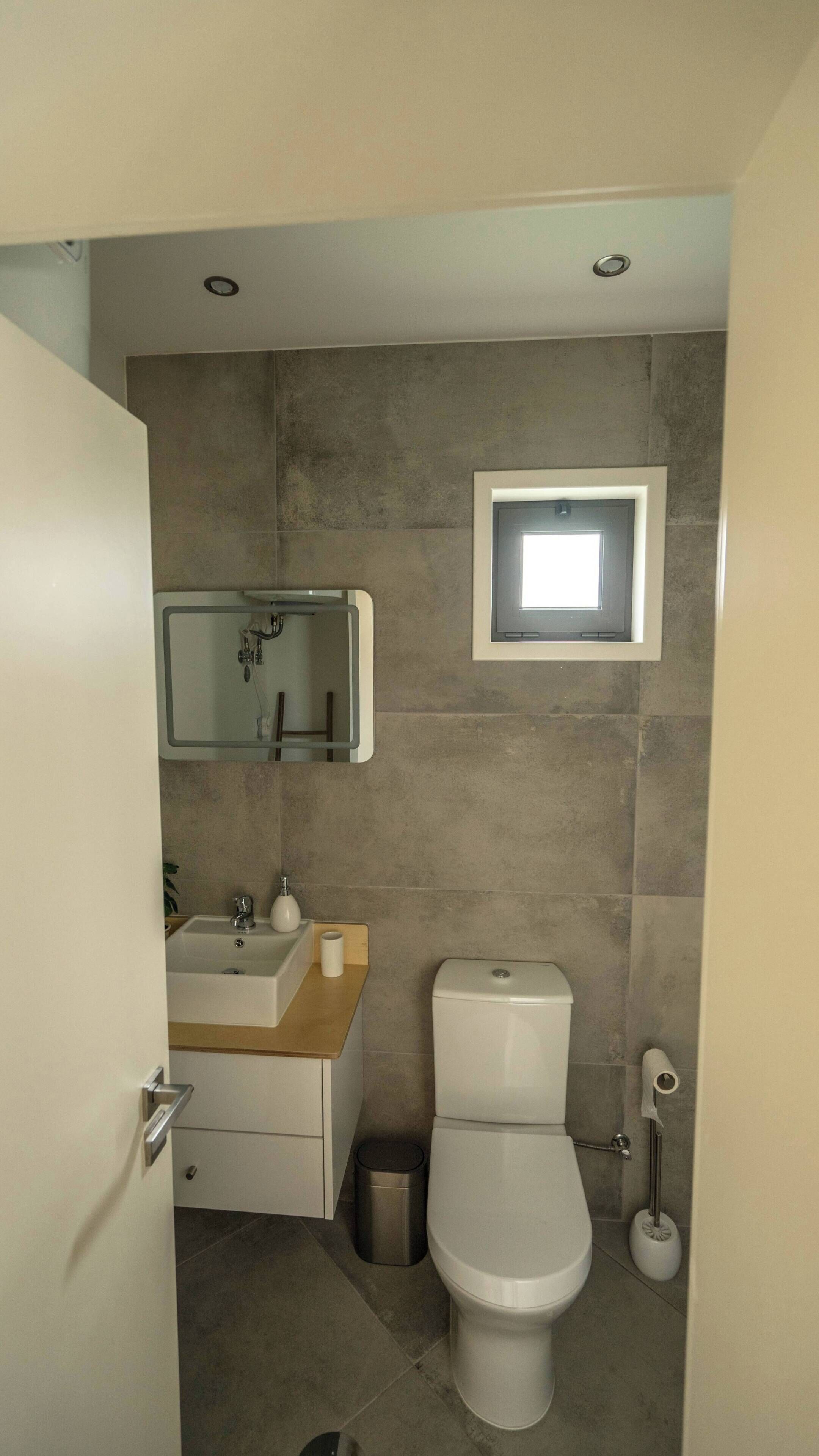 Un baño pequeño y funcional con lavamanos, espejo, inodoro y una ventana.