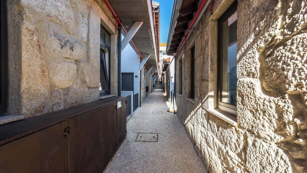 Um corredor estreito com paredes de pedra e janelas, que dá para uma área coberta com chão de azulejos.