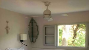 A bedroom with a bed, a ceiling fan, a window with a view of greenery, and a wall-mounted light fixture.