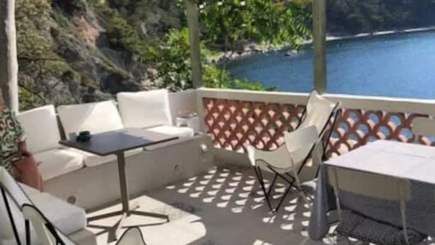 A covered outdoor seating area with a table and chairs, overlooking a body of water and greenery.