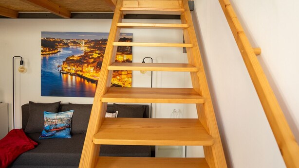 A modern interior with a wooden staircase, a sofa with cushions, a framed cityscape picture, and a small red object on the wall. - Ilha D’ouro House S. Vitor II (Porto)