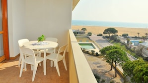 A balcony with a round table and chairs, overlooking a beach and a pool area. - Find Peace by the sea in our Welcoming Flat (San Michele al Tagliamento)