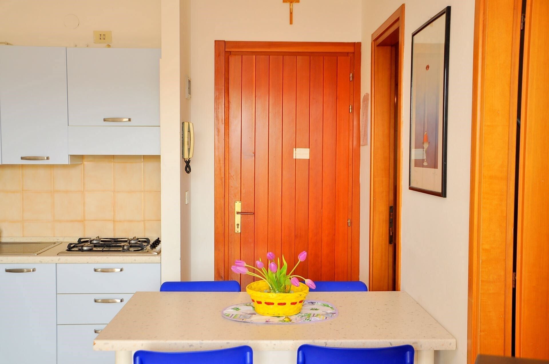 A small kitchen with white cabinets, a stove, and a dining table with blue chairs. There is a wooden door with a small window, and a framed picture on the wall.