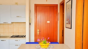 A small kitchen with white cabinets, a stove, and a dining table with blue chairs. There is a wooden door with a small window, and a framed picture on the wall. - Find Peace by the sea in our Welcoming Flat (San Michele al Tagliamento)