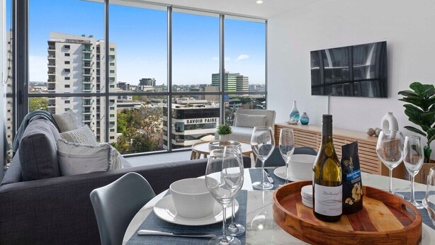 Un salon moderne avec un coin repas, de grandes fenêtres donnant sur la ville, un téléviseur à écran plat et une bouteille de vin posée sur la table.