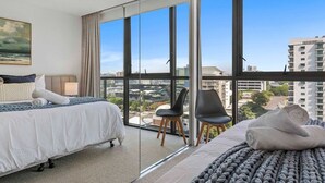 Une chambre d’hôtel moderne avec un grand lit, un lit plus petit, une chaise et une vue sur la ville grâce à de grandes fenêtres.