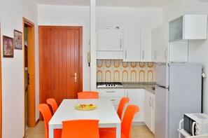 Eine kleine Küche mit weißen Schränken, einer Holztür, einem Esstisch mit orangefarbenen Stühlen und einem kleinen Kühlschrank.