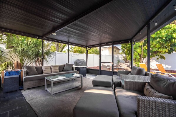 A covered outdoor seating area with a sofa, ottoman, and a glass coffee table.