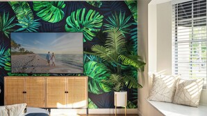 A room with a large TV, a rattan cabinet, a window seat with cushions, and a tropical leaf wallpaper.