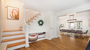 A staircase with wooden steps and a white railing, a dining area with a white table and chairs, and a kitchen area with a chandelier.