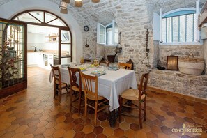 A rustic dining area with a stone wall, wooden chairs, a round table, and a view of the kitchen. - Château d'Aiguèze (Aiguèze)