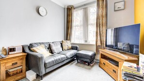 A modern living room with a grey leather sofa, a wooden TV stand, a radio, and a bookshelf. - GuestReady - Tranquility near Shepherd's Bush (London)