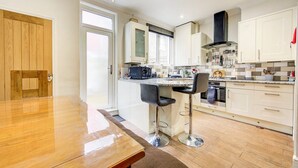 A modern kitchen with a wooden dining table, two bar stools, and a large window. - GuestReady - Tranquility near Shepherd's Bush (London)