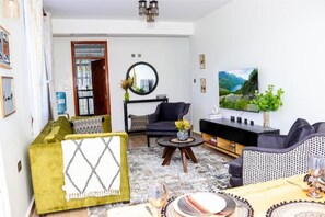 A living room with a sofa, armchairs, a coffee table, and a dining area with a table set for two. - Deluxe king suite (Nairobi)