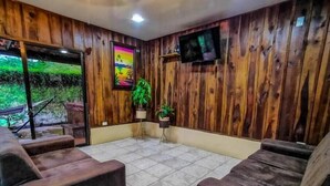A room with wooden panel walls, a TV mounted on the wall, and a large window with a view of greenery outside. - Cabaña el Descanso Fortuna (La Fortuna)