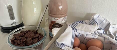 A kitchen counter with a blender, a glass jar of milk, a bottle of chocolate milk, bowls of cereal, and a basket of eggs.