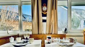 A dining area with two tables set for two, each with a white tablecloth, plates, cutlery, and glasses. A bottle of Nutella is on one table. The room has large windows with curtains, a clock on the wall, and a view of a building and trees outside.