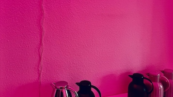 Een neonbord met de tekst 'ERSTMALE KAFFEE' op een roze muur, met verschillende koffiepotten op een plank eronder.