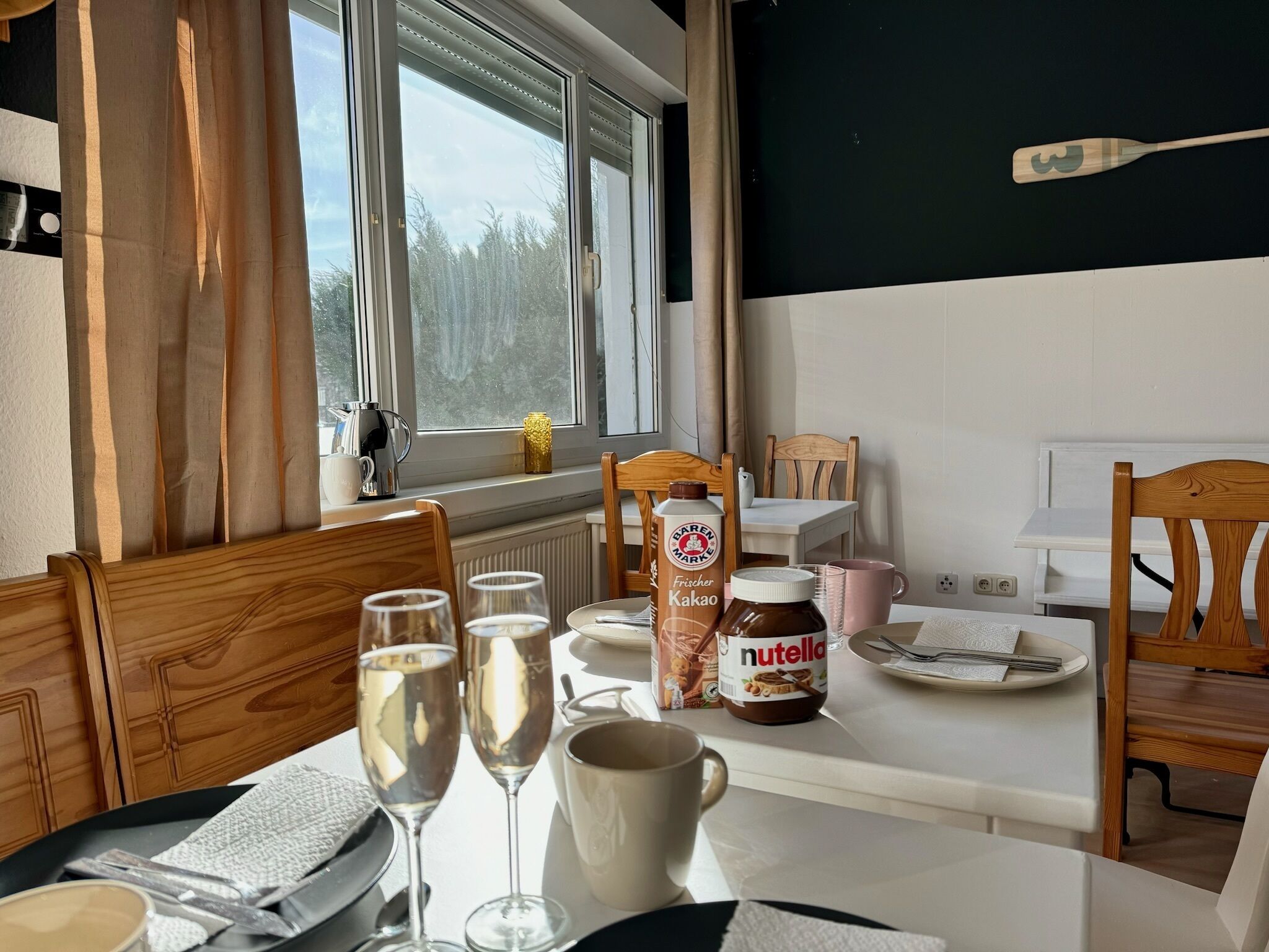 Uma área de refeições com uma mesa posta para dois, exibindo uma garrafa de Karen Marie Frischer Kakao, um pote de Nutella e dois copos de água.