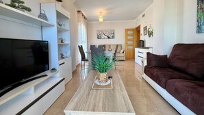 A modern living room with a flat-screen TV, a wooden coffee table, a brown sofa, and a dining area with a table and chairs. - Alq 119 Porto Azul 12 Apartment (Calpe)