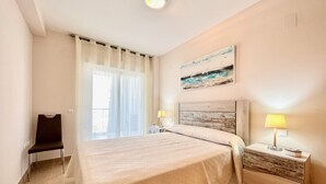 A hotel room with a large bed, a chair, a nightstand with a lamp, and a painting on the wall. - Alq 119 Porto Azul 12 Apartment (Calpe)