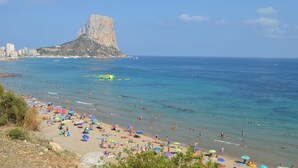 A beach with people swimming and sunbathing, a large rock formation in the background, and a clear blue sky. - Alq 119 Porto Azul 12 Apartment (Calpe)