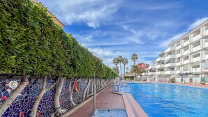 Una piscina con un muro de mosaico de azulejos, un paseo con baldosas rojas y un edificio con balcones al fondo.