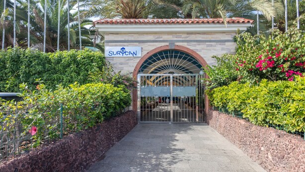Una entrada protegida por un portón con un letrero que dice “SURYCAN” y un muro de piedra adornado con follaje a ambos lados.