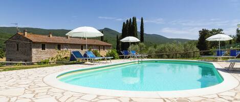 A swimming pool with blue lounge chairs and white umbrellas, surrounded by stone tiles and a stone building with a tiled roof.
