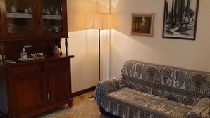 Uma sala de estar com um sofá estampado, um aparador de madeira com portas de vidro, um abajur e quadros na parede.
