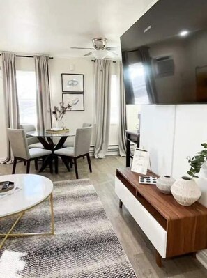 Una sala de estar moderna con comedor, televisor de pantalla plana, ventilador de techo y un ventanal con cortinas.