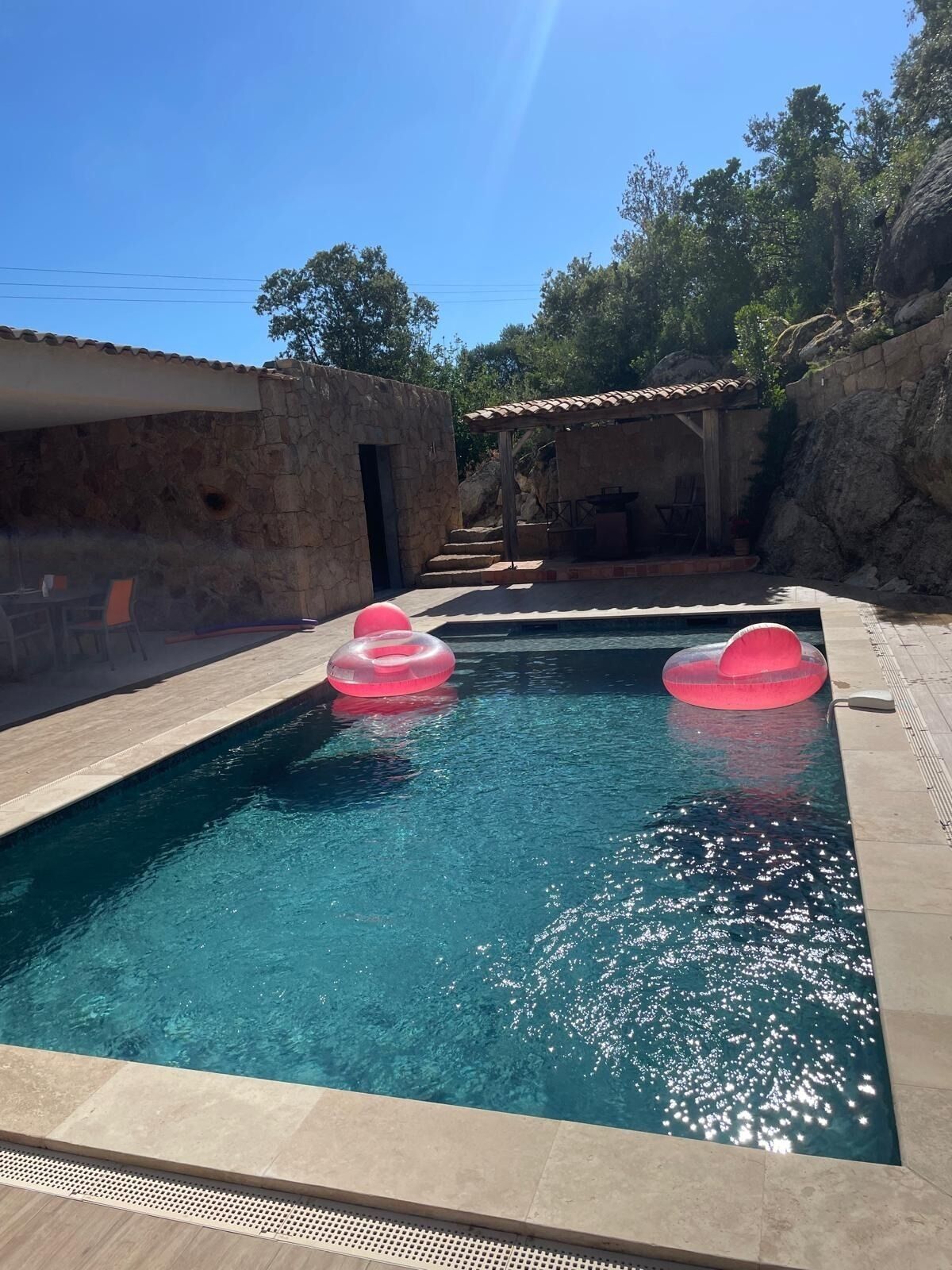 A swimming pool with two pink inflatable rings, stone walls, and a covered seating area.