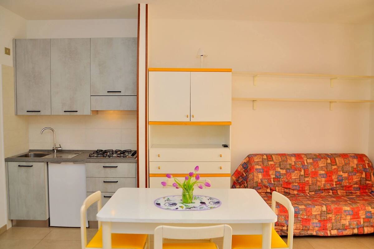 A compact kitchen with a gas stove, sink, and cabinets. There is a small dining table with chairs and a colorful cushion.