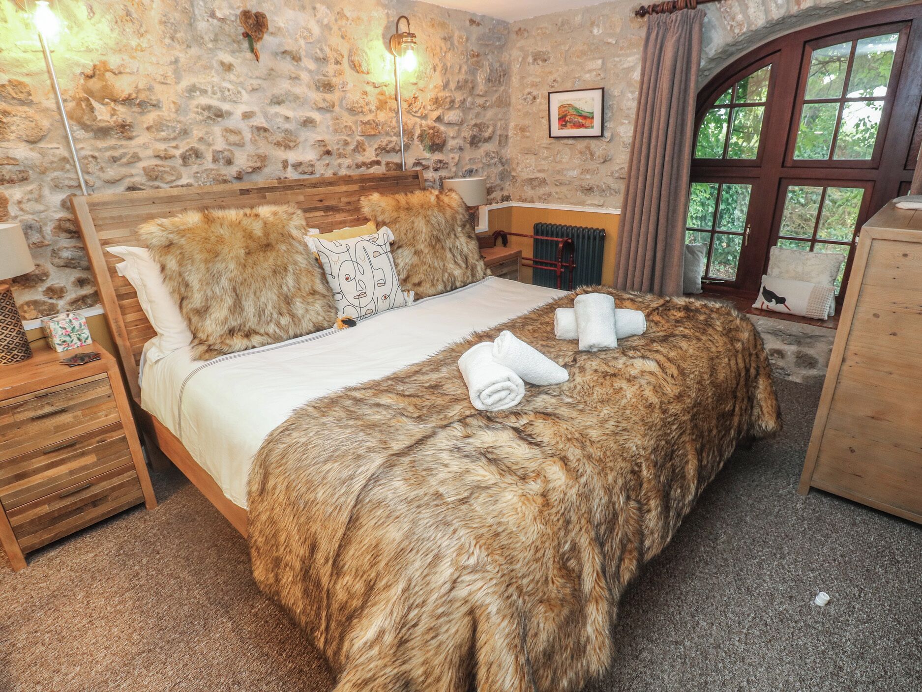 A bedroom with a stone wall, a bed with a fur throw, a wooden dresser, and a window with curtains.