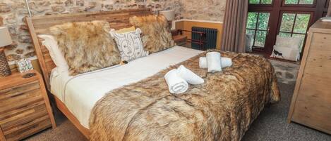 Een slaapkamer met een stenen muur, een bed met een bont plaid, een houten dressoir en een raam met gordijnen.