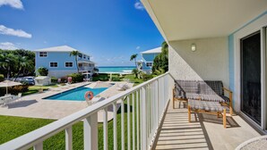 A balcony with a pool, lounge chairs, and a view of the ocean and neighboring buildings. - Beautiful beachfront apartment on Seven Mile Beach! (George Town)