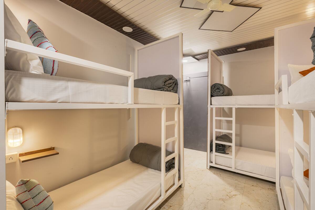 Une chambre de type dortoir, avec des lits superposés, une échelle, une étagère et un luminaire fixé au mur.